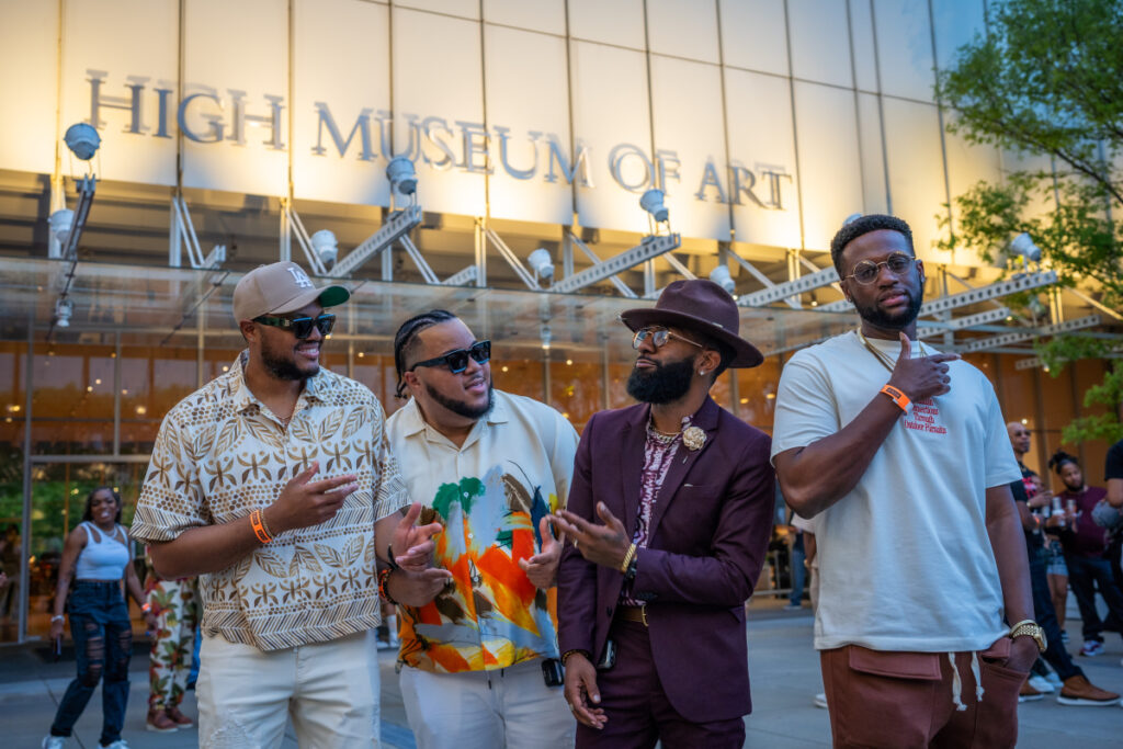 Four stylish men stand outside the High Museum of Art, smiling and talking. They wear colorful, trendy outfits and sunglasses, with the museum’s glass facade and logo visible in the background.