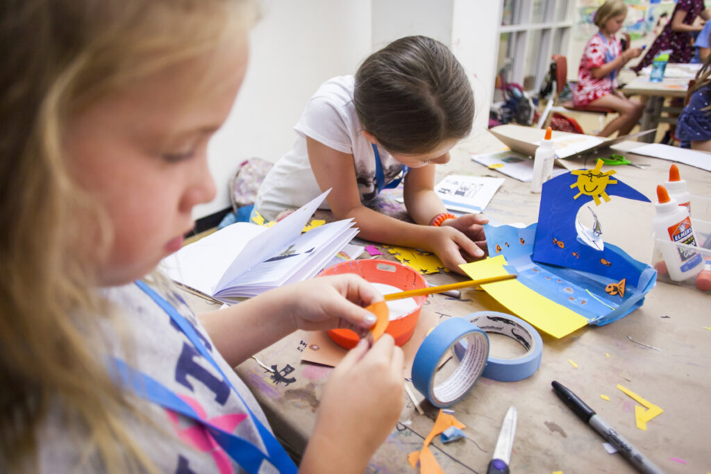 Two young children sit at a table crafting colorful paper projects with scissors, tape, and glue. One child focuses on cutting orange paper, while the other assembles a blue pop-up card with sun and star shapes.